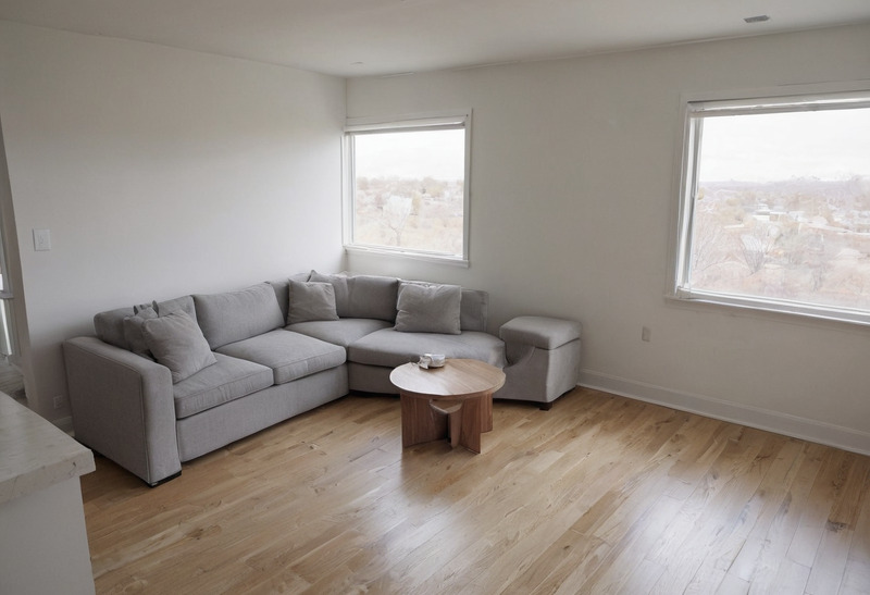 An open-plan living area with white walls, two windows, a light oak floor, a grey sectional sofa with an ottoman and a small round table, and clear floor in front of them.
