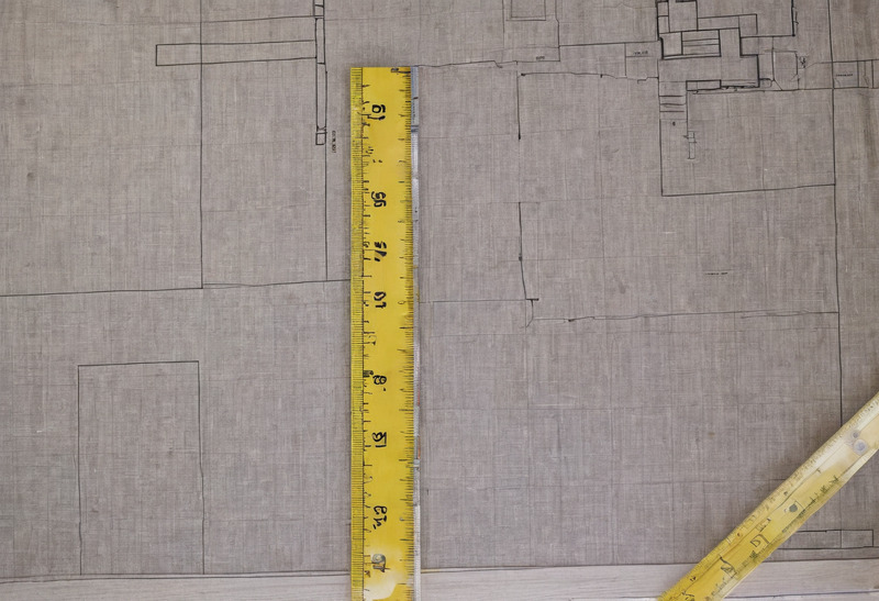 A printed floor plan seen from directly above with a yellow folding rule laid across it and the corner of a second rule at the edge of the sheet.