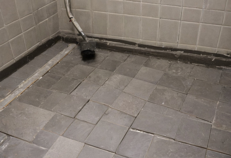 A floor of large grey square tiles newly laid, the perimeter still open to the adhesive bed and battens, with a vacuum hose reaching down into the corner.