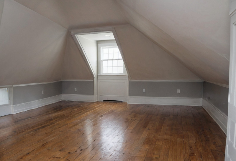A finished attic room with ceilings sloping to low knee walls on both sides, a dormer holding a double-hung window, a chair rail, and an old pine plank floor.